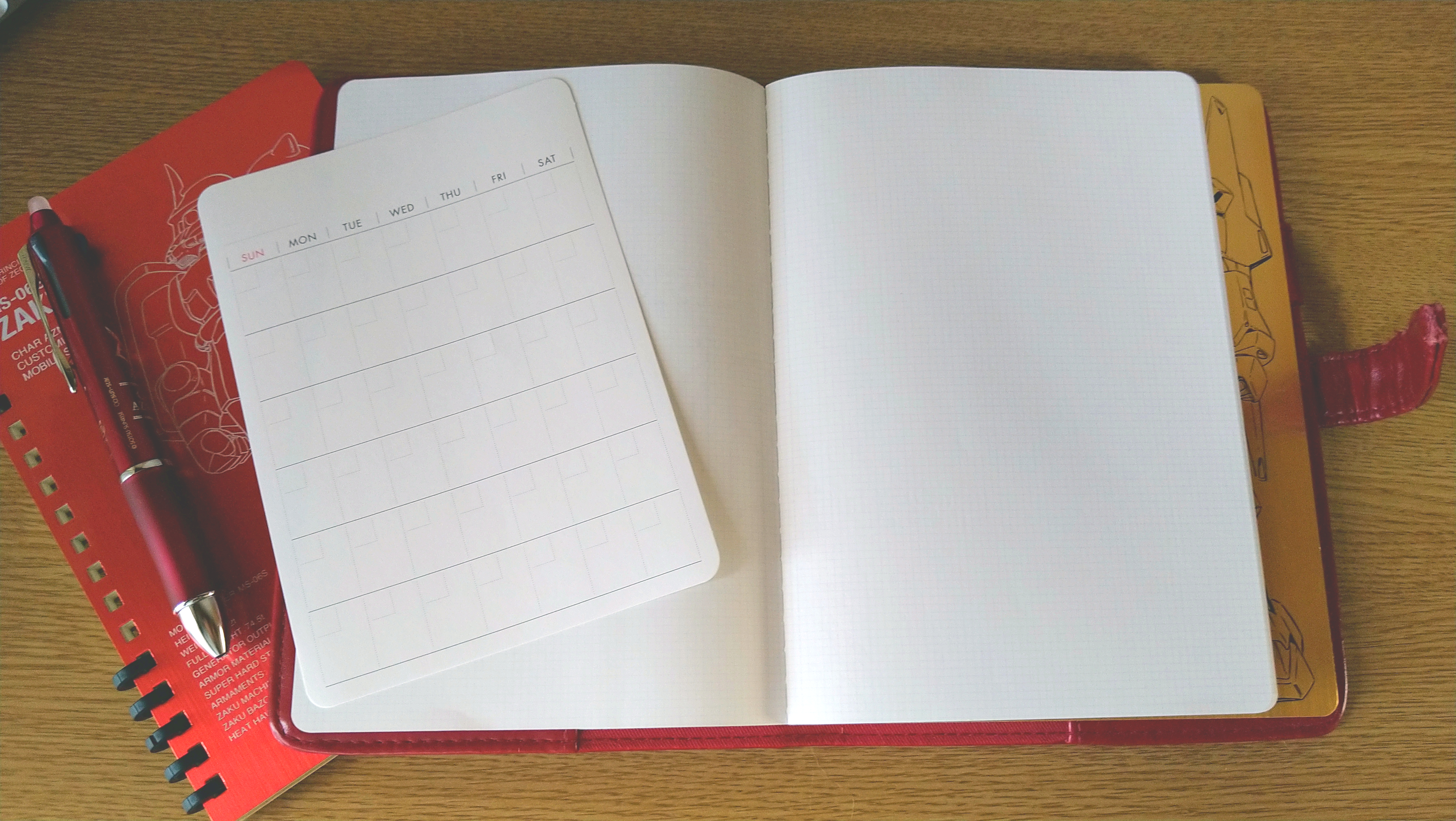 赤いノートカバーにセットされた真っ白な方眼ノートの見開き。左側のページには日付を書き込んで使えるフリータイプの月間カレンダーカードが置かれている。背景にはシャア専用ザクIIのノートと赤いボールペンが写っている